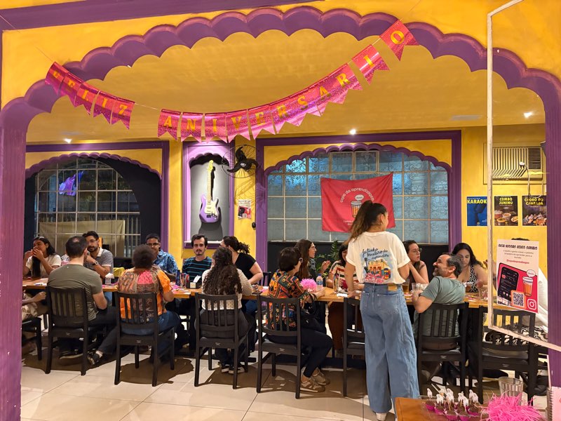 Salão de boteco amarelo e roxo com bandeirinhas de festa e uma mesa comprida cheia de participantes conversando.
