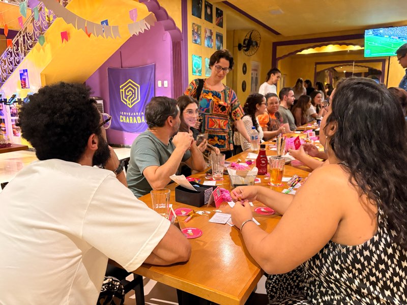 Pessoas em volta de uma mesa de madeira com bolachas de chope rosas e cartinhas do Copo espalhadas.