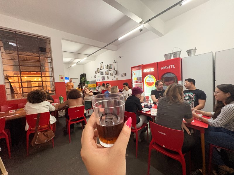 Mão segurando um copo americano com a marca do Copo da Aprendizagem, com o bar cheio ao fundo.