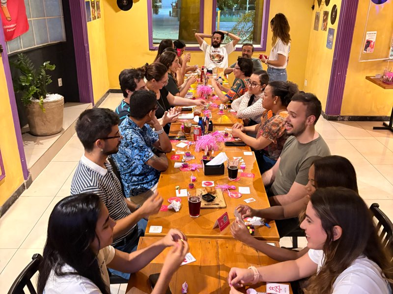Mesa comprida vista do alto, com participantes dos dois lados e enfeites cor-de-rosa no centro.