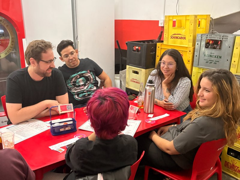 Roda de conversa em volta de uma mesa vermelha, com cartas e materiais do Copo sobre o tampo.