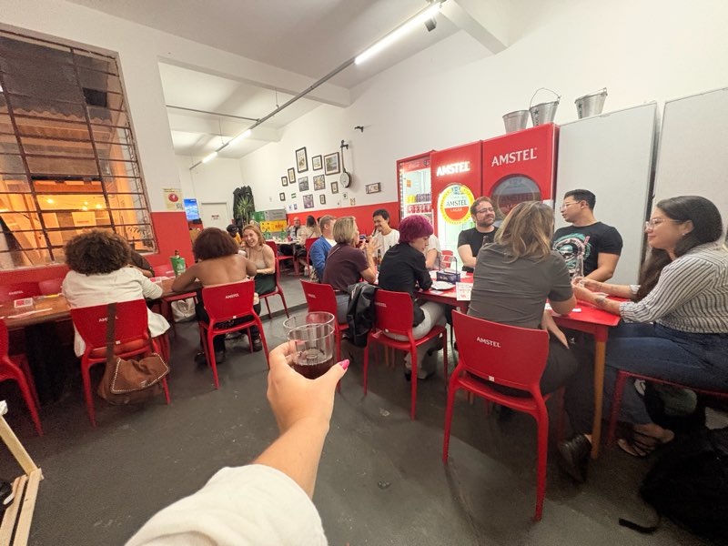 Copo levantado em primeiro plano com o salão do bar cheio de participantes ao fundo.